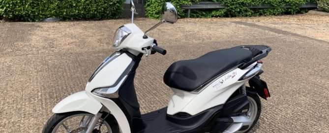 White piaggio liberty parked on the stand outside a building with a green hedge in the bcackground.