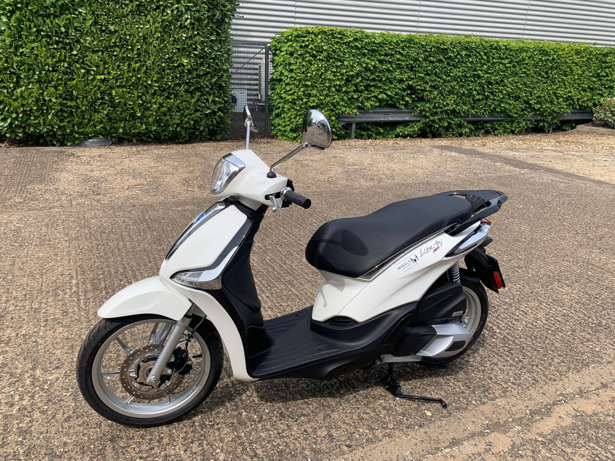 White piaggio liberty parked on the stand outside a building with a green hedge in the bcackground.