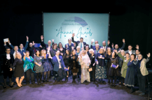 Large group of people stand on stage at the Royal Theatre with their arms in the air to celebrate Northamptonshire Community Foundation's Annual Awards