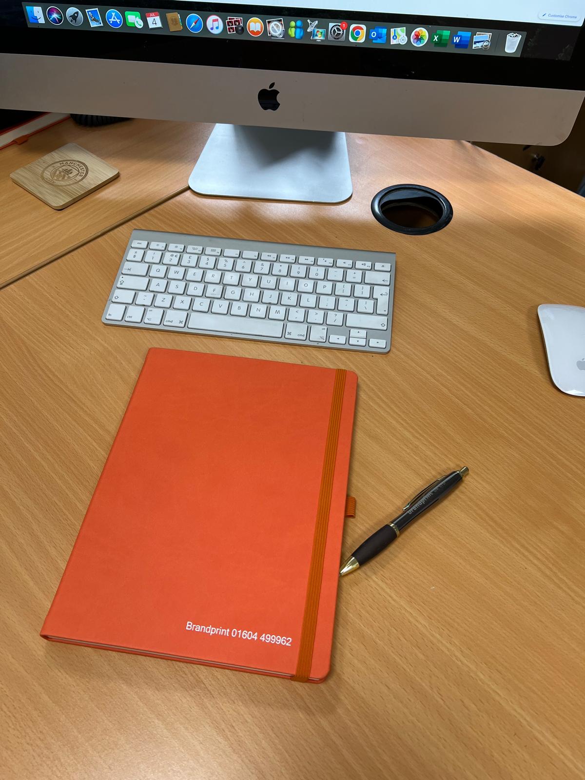 Orange 2026 branded diary on a wooden desk with pen and computer screen